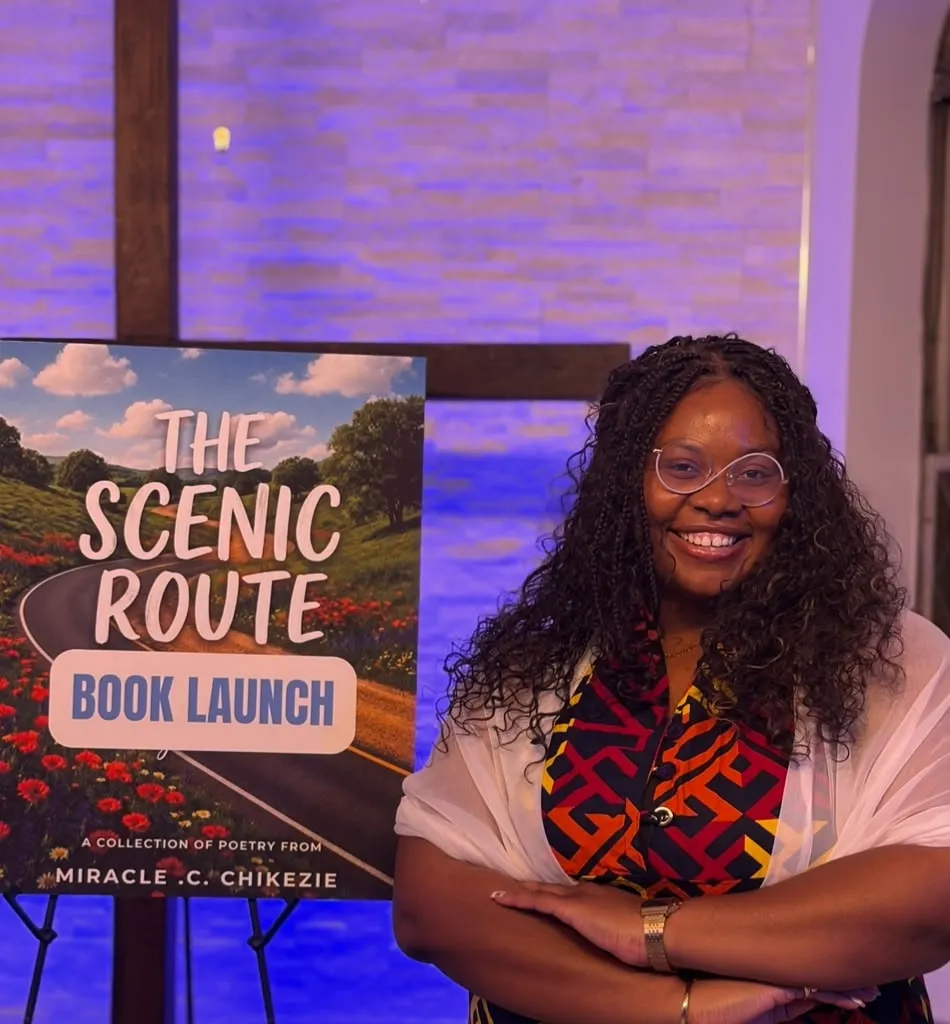 Miracle smiling beside an easel poster for The Scenic Route book launch.
